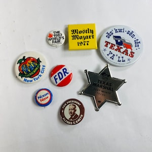 May include: A collection of vintage button pins featuring various designs, including a red, white, and blue pin with the text "Texas Ya'll", a yellow pin with the text "Mostly Mozart 1977", a red and white pin with the text "FDR", a blue and white pin with the text "Nixon Now", a brown pin with the text "For President Alfred E. Smith", a red and white pin with the text "The 1982 World's Fair", and a red and white pin with the text "New York City".