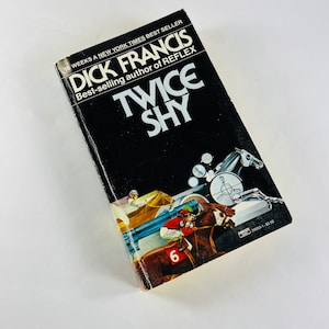 May include: A black paperback book with the title "Twice Shy" by Dick Francis, a New York Times best-selling author. The cover features an illustration of a horse race.