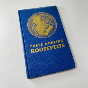 May include: A blue hardcover book titled "These Amazing Roosevelts." The book features a gold-coloured portrait of a man in a circle. The text and portrait are in gold. The book is on a white surface.