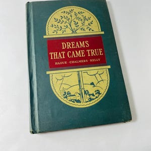 May include: A vintage book titled "Dreams That Came True" with a dark teal cover. The title is in red, with gold illustrations of a tree and a village. The book's cover shows signs of wear, indicating its age.