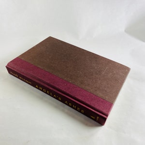 May include: A brown hardback book with a maroon spine. The spine has gold lettering that reads "Frank McCourt Angela's Ashes".