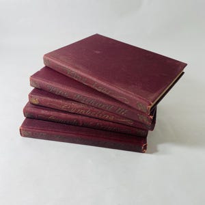 May include: A stack of five antique books with burgundy covers. The books are arranged in a slightly staggered pile, with the titles barely visible on the spines. The covers have a textured appearance, and the edges show signs of wear.