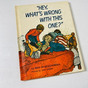 May include: A vintage children's book titled "Hey, What's Wrong With This One?" by Maia Wojciechowska with illustrations by Joan Sandin. The cover features a colorful illustration of three children on a bed.