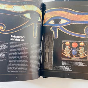 May include: Open book page featuring illustrations of ancient Egyptian artifacts. The page displays a detailed depiction of the Eye of Horus, with blue, gold, and black colors. Text on the page describes the God of the Sun and the significance of the eye.