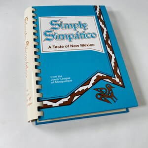 May include: A turquoise cookbook titled "Simply Simpático: A Taste of New Mexico" with white and brown geometric designs. The spiral-bound book is from the Junior League of Albuquerque.