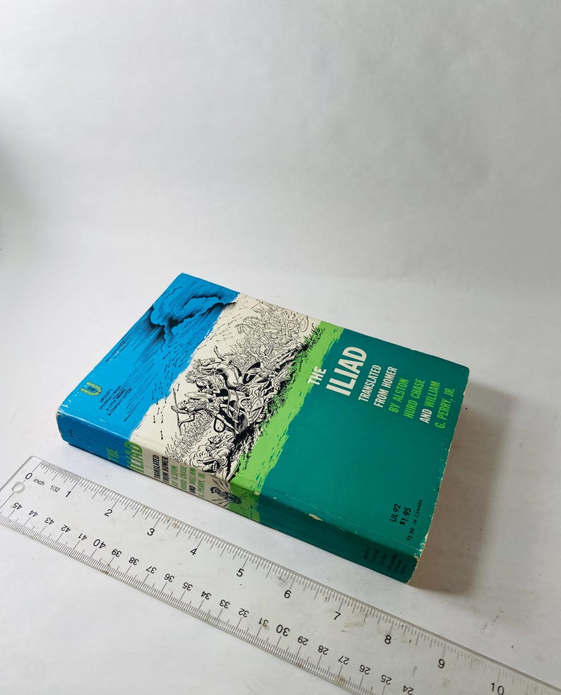 K&ouml;nnte beinhalten: Ein gebundenes Buch mit dem Titel "The Iliad" mit einem t&uuml;rkis-gr&uuml;nen Einband. Der Einband zeigt Illustrationen und Text, einschlie&szlig;lich der Namen der &Uuml;bersetzer. Ein Lineal liegt neben dem Buch.