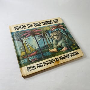 May include: A hardcover book titled Where the Wild Things Are, Story and Pictures by Maurice Sendak. The cover features a color illustration of a large, horned creature sitting in a forest near a sailboat on water, with aged, yellowed edges.