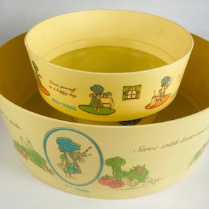 May include: Two yellow plastic mixing bowls, one nested inside the other. The bowls feature illustrations of a girl and vegetables, with text that reads "Treat yourself to a happy day" and "Serve with love and a smile."