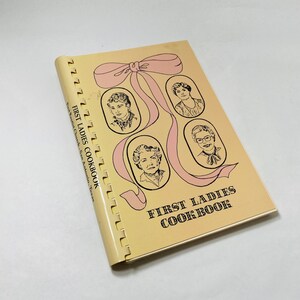 Könnte beinhalten: Ein gelbes Kochbuch mit einer rosa Schleife und Illustrationen von vier Frauen. Der Titel des Buches lautet "First Ladies Cookbook".