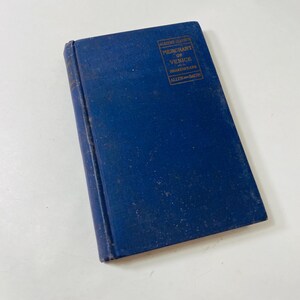 May include: A vintage hardcover book with a dark blue cover. The title "Merchant of Venice" is visible in gold lettering, along with the author's name and publisher. The book is slightly worn, suggesting age and use.