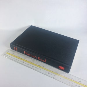 May include: A black hardcover book with the title "Destiny's Road" in red lettering on the spine. The book is lying on a white surface.