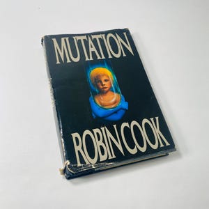 May include: A black hardcover book with the title "Mutation" in white letters. The cover features a child with blonde hair in a blue tube. The author's name, "Robin Cook", is printed in white letters below the image.