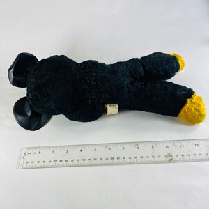 May include: Black plush toy with yellow feet and black ears. The toy is lying on a white surface.