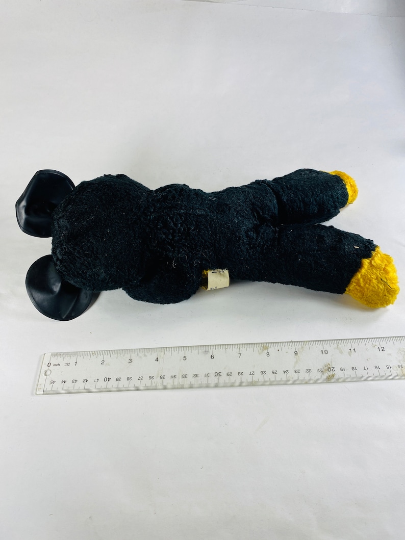 May include: Black plush toy with yellow feet and black ears. The toy is lying on a white surface.