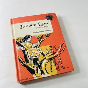 May include: Vintage book titled "Juliette Low Girl Scout" by Helen Boyd Higgins. The cover is orange with an illustration of a girl in a tree and two boys. The book is illustrated by Anne Fleur.