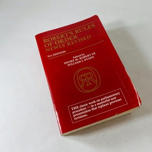 May include: A red book titled "Robert's Rules of Order Newly Revised" with gold lettering. The book's cover includes the text "9th Edition" and the names of the editors. A gold emblem is also visible.