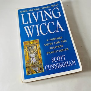 May include: A hardcover book titled "Living Wicca" with a blue cover and white text. The cover features the text "Over 200,000 Copies Sold!" and "A Further Guide for the Solitary Practitioner" by Scott Cunningham. An illustration of a woman is on the cover.