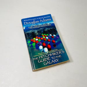 Könnte beinhalten: Ein Taschenbuch mit dem Titel "The Hitchhiker's Guide to the Galaxy" von Douglas Adams. Das Cover zeigt farbige Kugeln und Text. Das Buch gehört zur Hitchhiker-Trilogie.
