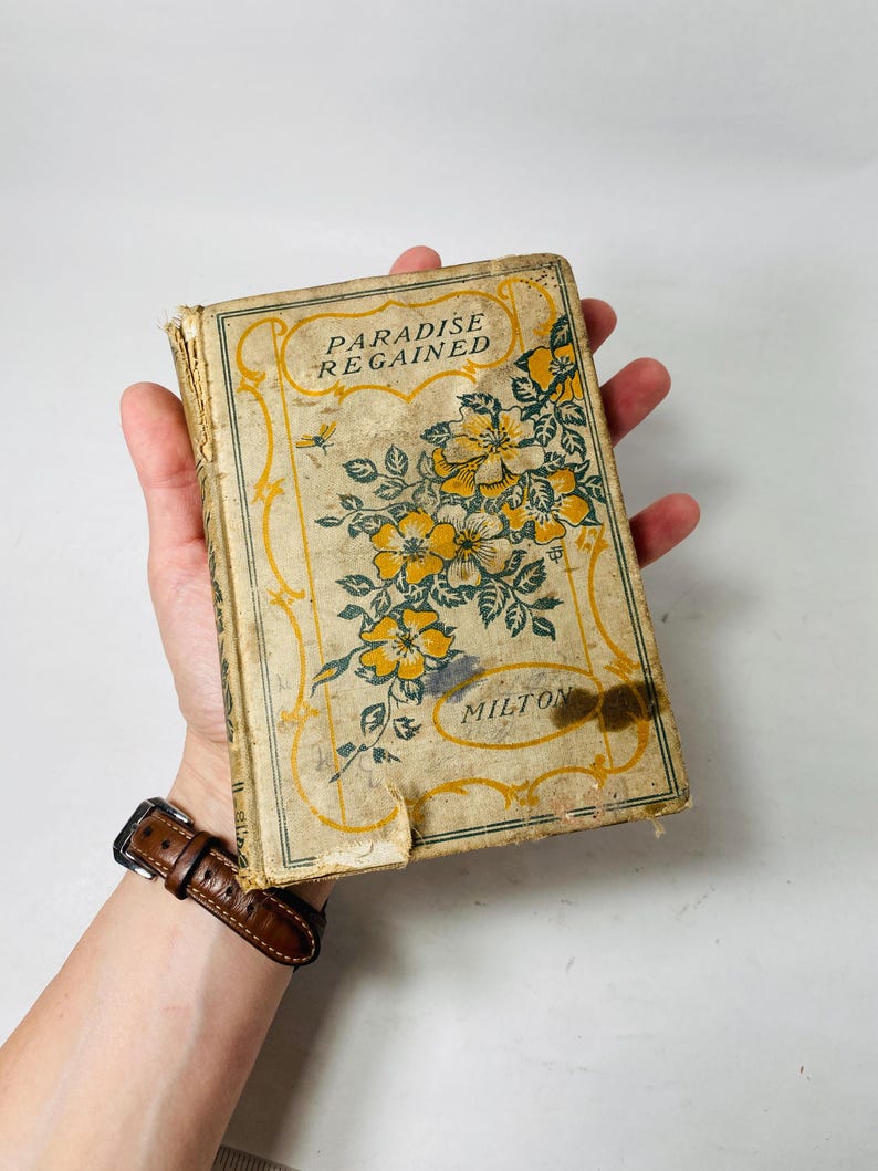 May include: An antique book titled "Paradise Regained" by Milton. The cover is beige with yellow floral illustrations and text. The book shows signs of wear and tear, with a visible stain on the cover.