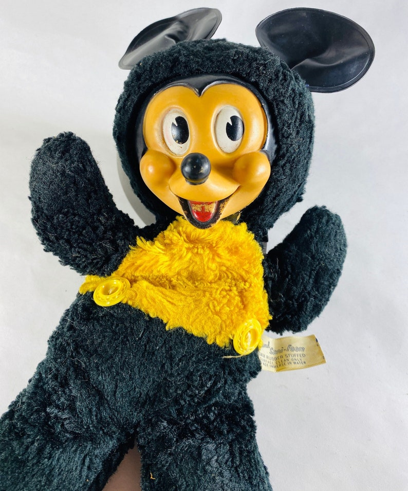 May include: Vintage black and yellow plush Mickey Mouse toy with a plastic face and button eyes. The toy is wearing a yellow vest with two yellow buttons. The tag reads "Hand Sewn - Foam Rubber Stuffed - Washable - Clean with Mild Soap & Water - Made in U.S.A."
