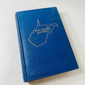 May include: A blue book titled "West Virginia Blue Book" with gold lettering and a gold outline of the state. The book has a textured cover and is slightly angled, showcasing its title and design.
