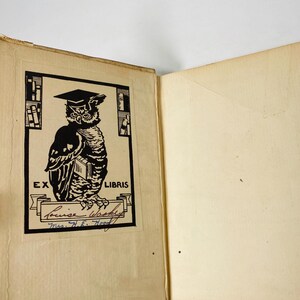 May include: Open book with a black and white illustration of an owl wearing a graduation cap. The owl is holding a book and is inside a rectangular frame with the words "EX LIBRIS" at the top. Below the owl is a banner with the name "Louise Washke" and "Mrs. H.E. Hood" written below.