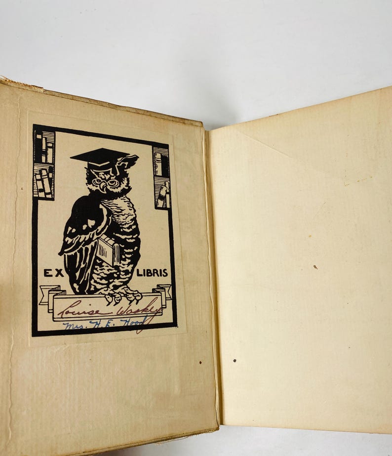 May include: Open book with a black and white illustration of an owl wearing a graduation cap. The owl is holding a book and is inside a rectangular frame with the words "EX LIBRIS" at the top. Below the owl is a banner with the name "Louise Washke" and "Mrs. H.E. Hood" written below.