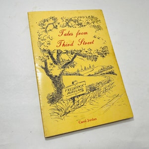 May include: A vintage book titled "Tales from Third Street" by Carol Jordan. The cover is yellow with a black and red illustration of a tree, a road sign for Frederick, Maryland, and a rural landscape.