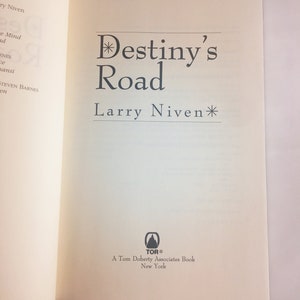 May include: A white book with the title "Destiny's Road" by Larry Niven. The book cover features a black asterisk before and after the title. The book is published by Tor Books, a Tom Doherty Associates book.