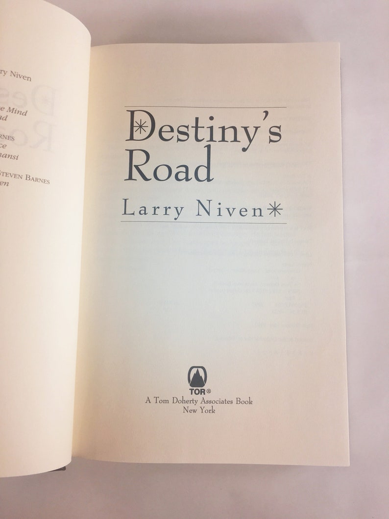 May include: A white book with the title "Destiny's Road" by Larry Niven. The book cover features a black asterisk before and after the title. The book is published by Tor Books, a Tom Doherty Associates book.