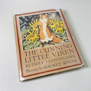 May include: A vintage book cover with a fox illustration. The title is "The Cunning Little Vixen" by Rudolf Tesnohlıdek. The cover features a fox standing in a field of sunflowers with a full moon in the background. The text "Pictures by Maurice Sendak" is printed below the title.