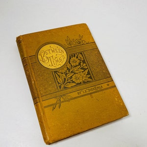 May include: A vintage book titled "Between Times" with a gold and brown cover. The cover features a circular design with the title in gold lettering, surrounded by floral and geometric patterns. The author's name, L.E. Diekenga, is printed at the bottom.