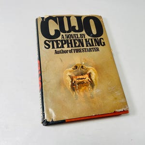 May include: A paperback book titled "CUJO" by Stephen King. The cover features a menacing dog's face with exposed teeth against a tan background. The book's title is in large, black, bold letters.