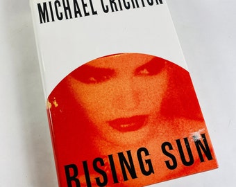 May include: A white hardcover book with the title "Rising Sun" in black text on a red background. The author, Michael Crichton, is printed in black text on the front cover.