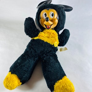 May include: Vintage black and yellow plush Mickey Mouse toy with a smiling face and button eyes. The toy has a yellow belly and yellow feet.