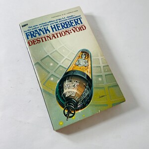 May include: A paperback book titled "DESTINATION: VOID" by Frank Herbert. The cover art features a black and gold spacecraft with a figure inside, set against a green and yellow background. The book is a science fiction classic.