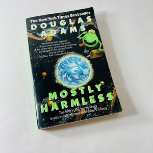 Könnte beinhalten: Ein gebundenes Buch mit dem Titel "Mostly Harmless" von Douglas Adams. Das Cover ist schwarz mit grünen und blauen Akzenten, mit einer Cartoon-Figur und einer Planetenillustration. Das Buch gehört zur Hitchhiker-Trilogie.