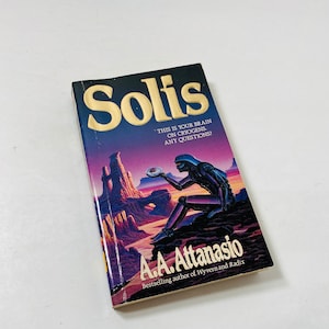May include: A paperback book cover with a gold title "Solis" and a futuristic illustration of a robot holding a glowing orb. The text on the cover reads "This is your brain on cryogens. Any questions?" and "A.A. Attanasio, Bestselling author of Wyvern and Radix."