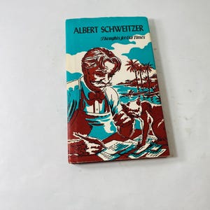 Könnte beinhalten: Ein Vintage-Buch mit dem Titel "ALBERT SCHWEITZER Thoughts for Our Times". Das Cover zeigt eine Illustration eines Mannes mit Schnurrbart, eines Hundes und einer tropischen Szene in Rot-, Blau- und Weißtönen. Das Buch liegt auf einer weißen Oberfläche.