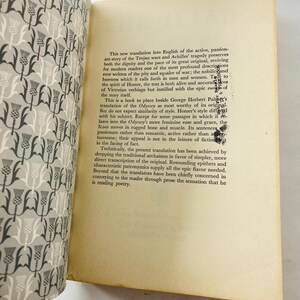 K&ouml;nnte beinhalten: Ein offenes Buch mit Text auf gealtertem Papier. Der Bucheinband zeigt ein sich wiederholendes Muster aus grauen und wei&szlig;en distelartigen Designs. Der Text beschreibt eine &Uuml;bersetzung der Ilias.