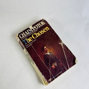 May include: A paperback book titled "The Chosen" by Chaim Potok, author of "Davita's Harp". The book cover features a portrait of a man wearing glasses and a dark sweater, holding a stack of books.