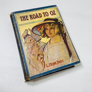 May include: A vintage hardback book titled "The Road to Oz" by L. Frank Baum. The cover features a colourful illustration of a young girl wearing a straw hat and a whimsical character holding a watering can.