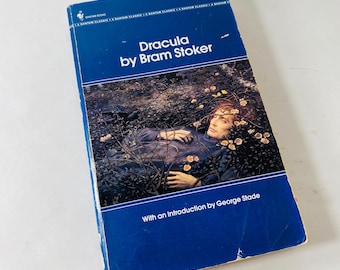 Puede incluir: Un libro de bolsillo vintage titulado "Dracula" de Bram Stoker. La portada es predominantemente azul con el título en blanco. La ilustración de la portada muestra a un hombre en la naturaleza. El libro incluye una introducción de George Slade.