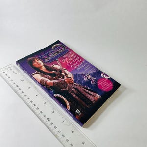 Pu&ograve; includere: Un libro tascabile intitolato Xena: All I Need to Know I Learned From the Warrior Princess, con un'illustrazione di Xena che impugna una spada. Un righello di plastica trasparente &egrave; posto accanto al libro, indicando una lunghezza di circa 20,3 cm.