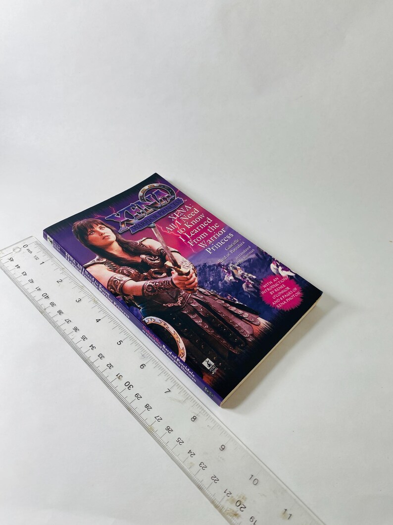 Pu&ograve; includere: Un libro tascabile intitolato Xena: All I Need to Know I Learned From the Warrior Princess, con un'illustrazione di Xena che impugna una spada. Un righello di plastica trasparente &egrave; posto accanto al libro, indicando una lunghezza di circa 20,3 cm.