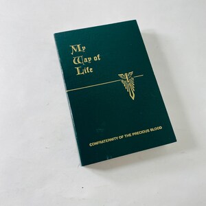 May include: A dark green book with gold lettering and a gold emblem. The text on the cover reads "My Way of Life" and "CONFRATERNITY OF THE PRECIOUS BLOOD". The book is rectangular and appears to be closed.