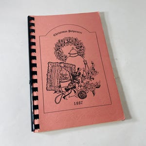 May include: A spiral-bound book with a salmon-colored cover featuring the title "Christmas Potpourri" and illustrations of a wreath, teapot, and other festive elements. The year "1987" is printed below the illustrations.