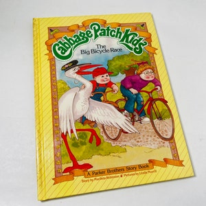 Könnte beinhalten: Ein Vintage-Kinderbuch mit dem Titel "Cabbage Patch Kids: The Big Bicycle Race". Das Cover ist gelb mit Illustrationen von Zeichentrickfiguren, die Fahrrad fahren. Das Buch ist ein Parker Brothers Story Book.