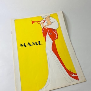 Peut inclure: Une impression rectangulaire avec un fond jaune vif et le mot MAME imprimé en noir. Une illustration stylisée montre une silhouette vêtue d'une longue robe blanche, d'une étole rouge et d'un col en fourrure blanche, tenant une trompette rouge.