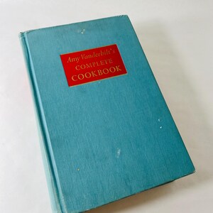 May include: A vintage cookbook titled "Amy Vanderbilt's Complete Cookbook." The book has a teal blue cover with a red rectangular label. The label features gold lettering. The book is slightly angled, showing its spine and cover.
